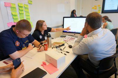 Three men and women sit at table during a discussion about artificial intelligence. One points to a computer screen with information displayed on it.